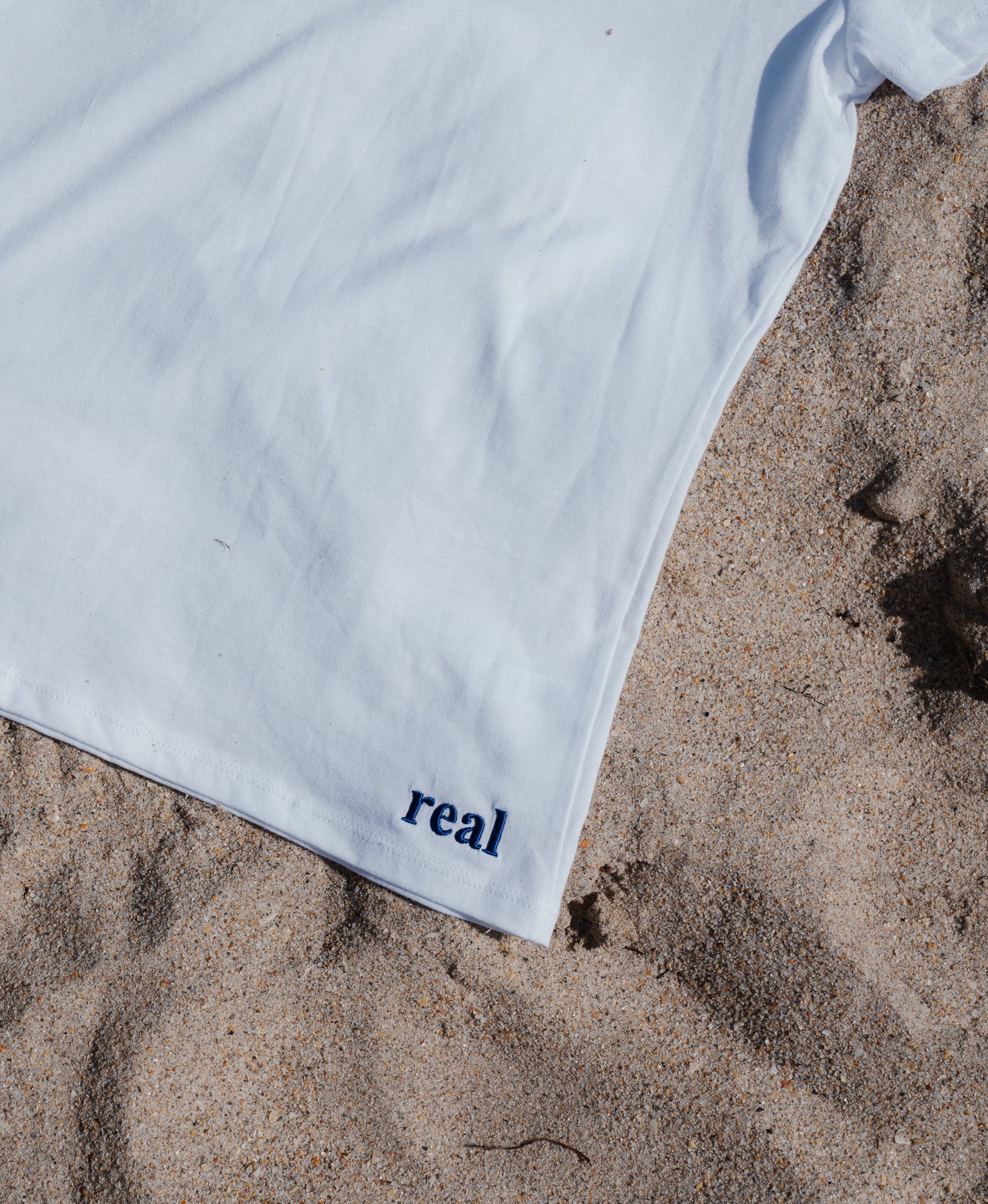 Women's White Tee with dark purple "real" embroidered on the left hip area | RaileyReal